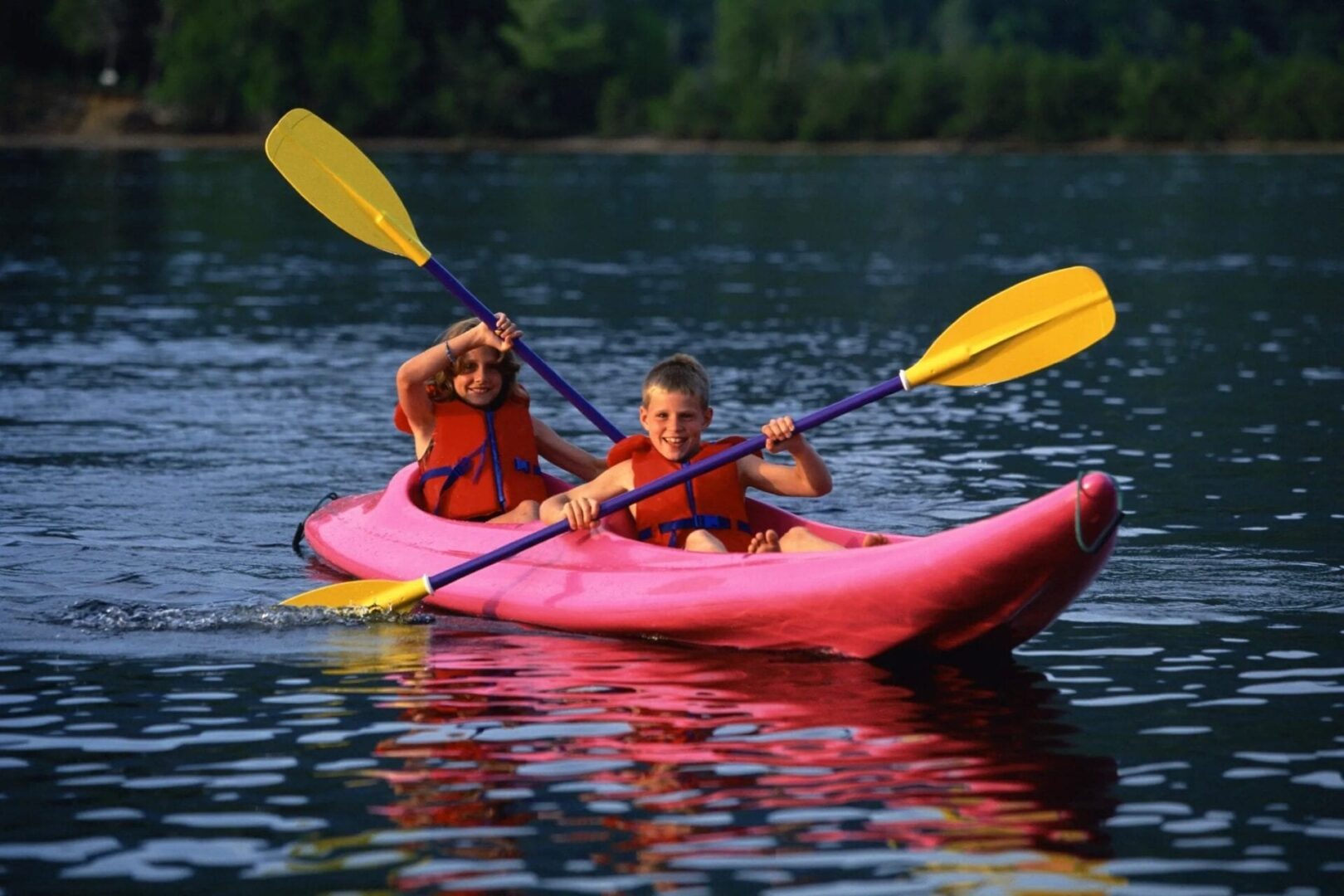 Two kids in a red kayak boat rowing using yellow and violet paddles